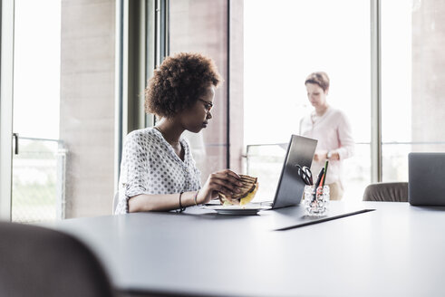 Businesswoman sitting at desk holding sandwich while looking at laptop - UUF008222