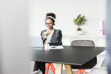 Businesswoman with headphones and beverage sitting at desk looking at laptop - UUF008230
