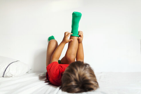 Rückenansicht eines kleinen Jungen, der auf dem Bett liegt und seine grünen Socken anzieht - VABF000715