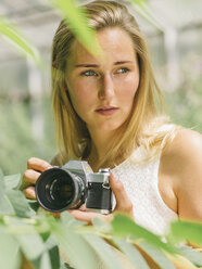 Young woman behind plant holding camera - MADF001050