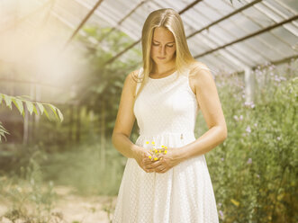 Blonde junge Frau in weißem Kleid steht im Gewächshaus und hält Blumen - MADF001062