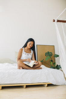 Junge Frau sitzt auf dem Bett und liest ein Buch - EBSF001556