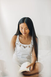 Junge Frau sitzt auf dem Bett und liest ein Buch - EBSF001557