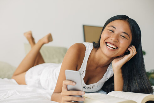 Lachende junge Frau im Bett liegend mit Handy in der Hand - EBSF001561