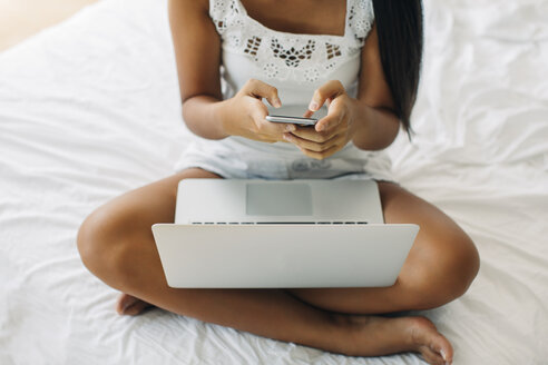 Junge Frau sitzt auf dem Bett und benutzt Handy und Laptop - EBSF001570