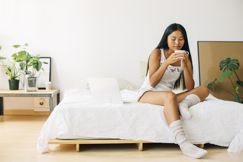 Junge Frau sitzt auf dem Bett und trinkt Kaffee - EBSF001572