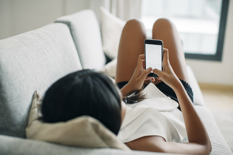 Junge Frau liegt auf der Couch und benutzt ein Mobiltelefon, lizenzfreies Stockfoto