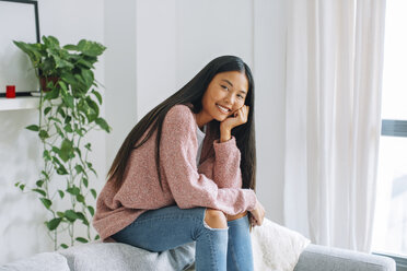 Smiling young woman sitting on backrest of couch at home - EBSF001612