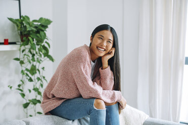 Laughing young woman sitting on backrest of couch at home - EBSF001613