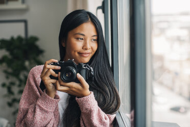Porträt einer lächelnden jungen Frau mit Kamera, die durch ein Fenster schaut - EBSF001617