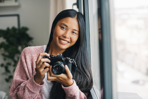 Porträt einer lächelnden jungen Frau mit Kamera in der Nähe des Fensters, lizenzfreies Stockfoto