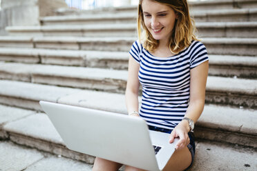 Lächelnde junge Frau, die auf einer Treppe sitzt und einen Laptop benutzt - GIOF001326