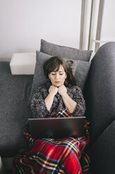 Junge Frau, zugedeckt mit einer Decke, auf der Couch liegend, mit Laptop - LCUF000026