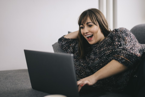 Erstaunte Frau auf der Couch liegend mit Blick auf den Laptop, lizenzfreies Stockfoto