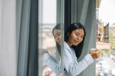 Young woman with cup of coffee leaning out of window - EBSF001628