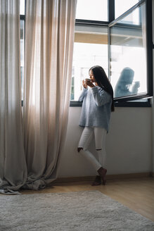 Junge Frau mit einer Tasse Kaffee vor einem offenen Fenster stehend - EBSF001629