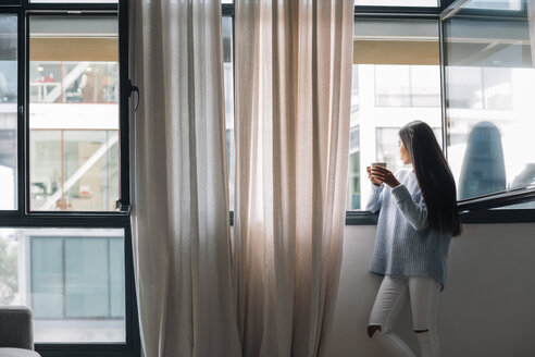 Junge Frau mit einer Tasse Kaffee schaut durch ein offenes Fenster - EBSF001630