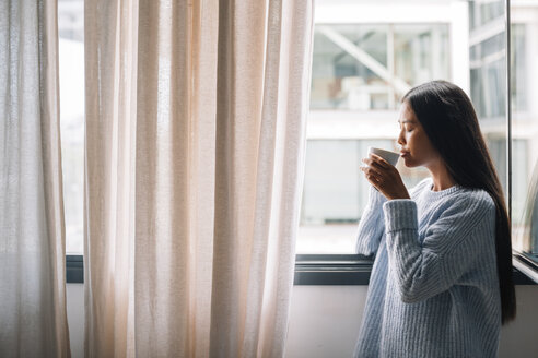 Junge Frau mit einer Tasse Kaffee vor einem offenen Fenster stehend - EBSF001631