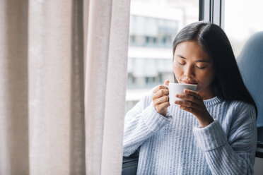 Junge Frau mit einer Tasse Kaffee vor einem offenen Fenster stehend - EBSF001633