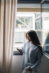Junge Frau mit einer Tasse Kaffee, die vor einem offenen Fenster steht und auf ihr Handy schaut - EBSF001634