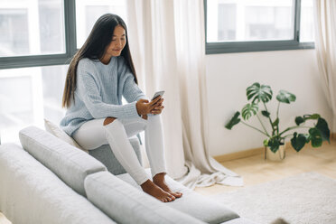 Junge Frau sitzt auf der Couch im Wohnzimmer und benutzt ein Smartphone - EBSF001635