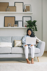 Lächelnde junge Frau sitzt zu Hause auf der Couch und benutzt einen Laptop - EBSF001647