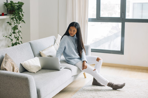 Junge Frau sitzt zu Hause auf der Couch und schaut auf den Laptop, lizenzfreies Stockfoto