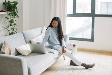 Young woman sitting on couch at home looking at laptop - EBSF001649