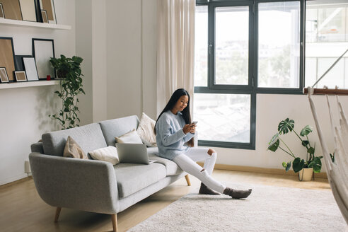 Young woman sitting on couch at home looking at smartphone - EBSF001650