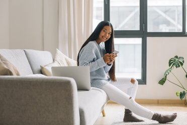 Smiling young woman sitting on couch with smartphone - EBSF001653
