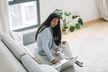 Young woman sitting on couch with smartphone looking at laptop - EBSF001655