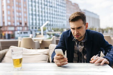 Junger Mann wartet in einem Straßencafé und schaut auf sein Handy - MGOF002119