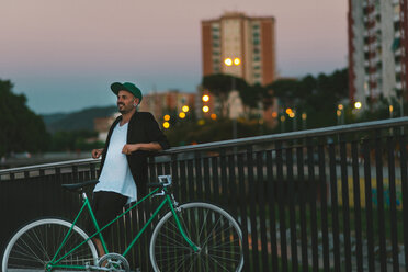 Lächelnder Mann mit Fahrrad, der sich in der Abenddämmerung an ein Geländer lehnt - SKCF000118