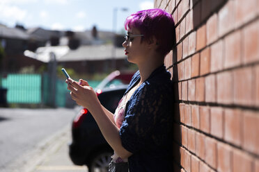 Junge Frau mit gefärbten Haaren lehnt an einer Backsteinmauer und schreibt eine SMS - BOYF000462