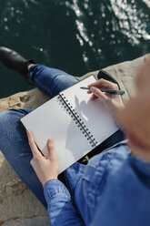 Junge Frau sitzt auf einem Steg und schreibt in ein Notizbuch - BOYF000493