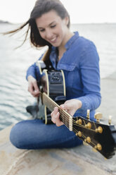Lächelnde junge Frau sitzt auf einem Steg und spielt Gitarre - BOYF000499