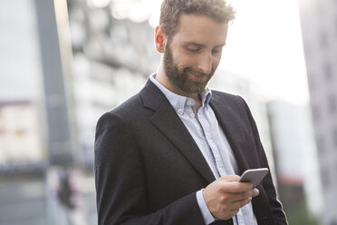 Smiling young businessman looking at cell phone outdoors - FKF001996