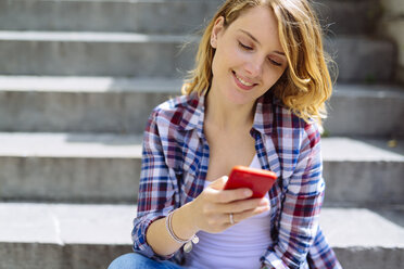 Porträt einer lächelnden jungen Frau, die auf einer Treppe sitzt und ein Smartphone benutzt - GIOF001358