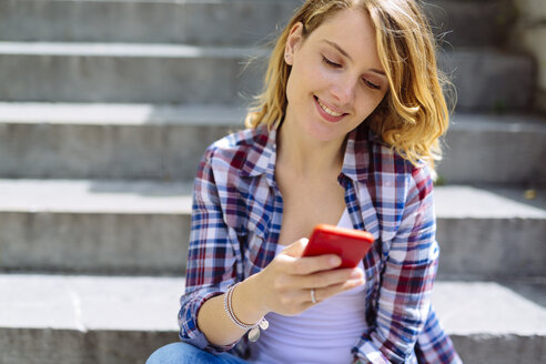 Porträt einer lächelnden jungen Frau, die auf einer Treppe sitzt und ein Smartphone benutzt - GIOF001358