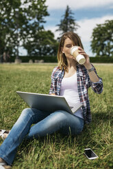 Junge Frau mit Laptop sitzt auf einer Wiese und trinkt einen Kaffee zum Mitnehmen - GIOF001364