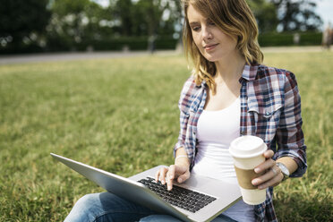 Junge Frau mit Kaffee zum Mitnehmen auf einer Wiese sitzend mit Laptop - GIOF001365