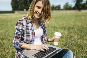 Lächelnde junge Frau mit Kaffee zum Mitnehmen, die auf einer Wiese sitzt und einen Laptop benutzt - GIOF001367