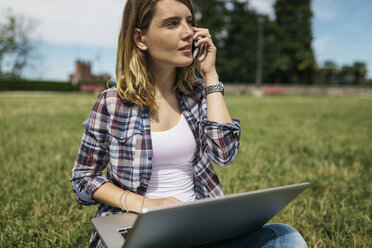 Junge Frau mit Laptop auf einer Wiese sitzend und mit Smartphone telefonierend - GIOF001368