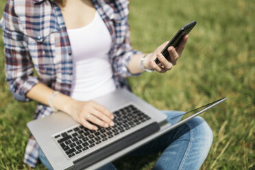 Junge Frau sitzt auf einer Wiese und benutzt Laptop und Smartphone, Teilansicht - GIOF001369