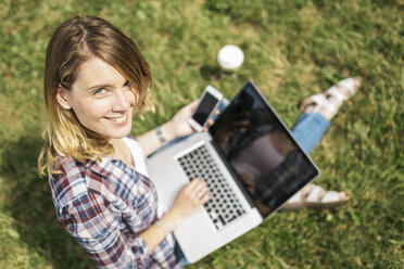 Smiling young woman sitting on a meadow using laptop and smartphone - GIOF001370