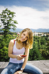 Smiling young woman with smartphone sitting on a wall - GIOF001375