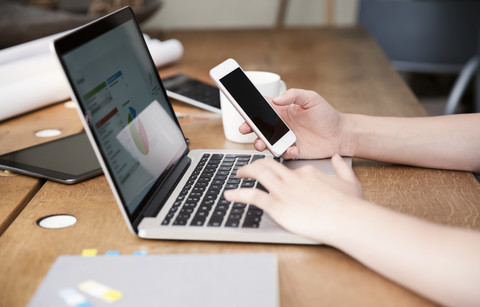 Frau am Schreibtisch mit Laptop und Smartphone arbeitet an einer Berechnung, lizenzfreies Stockfoto