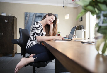 Smiling woman sitting on health chair at desk using laptop - REAF000098