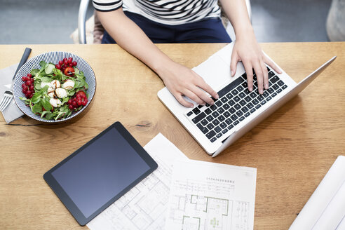 Woman at desk using laptop next to construction plan and salad - REAF000111