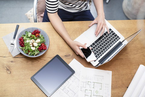 Woman at desk using laptop and cell phone next to construction plan and salad stock photo